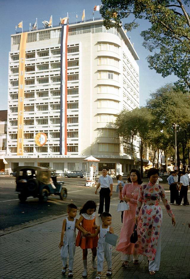 #38 Women dressed in ‘Ao Dai’ stroll down the street with their children in Saigon, 1961