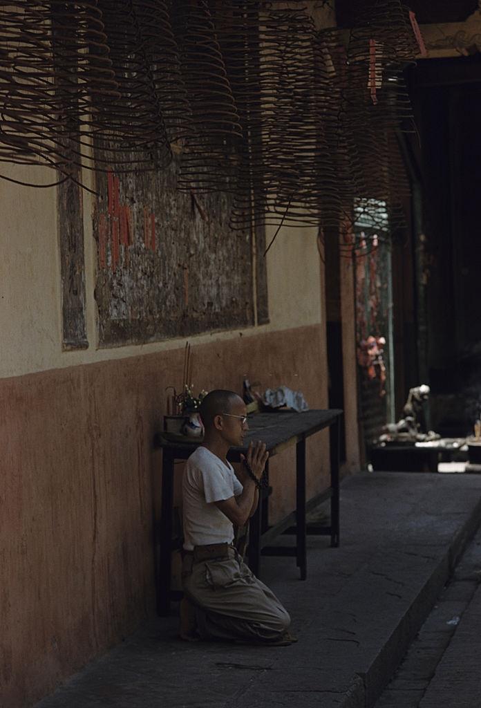 #42 A man kneeling on a sidewalk, prays with folded hands, holding a Buddhist Mâlâ, 1961.