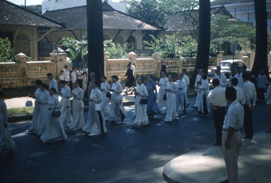 #44 Female mourners, wearing white ao dai, walking on a tree-lined street during Colonel Hoang Thuy Nam’s funeral procession, Saigon, 1961.