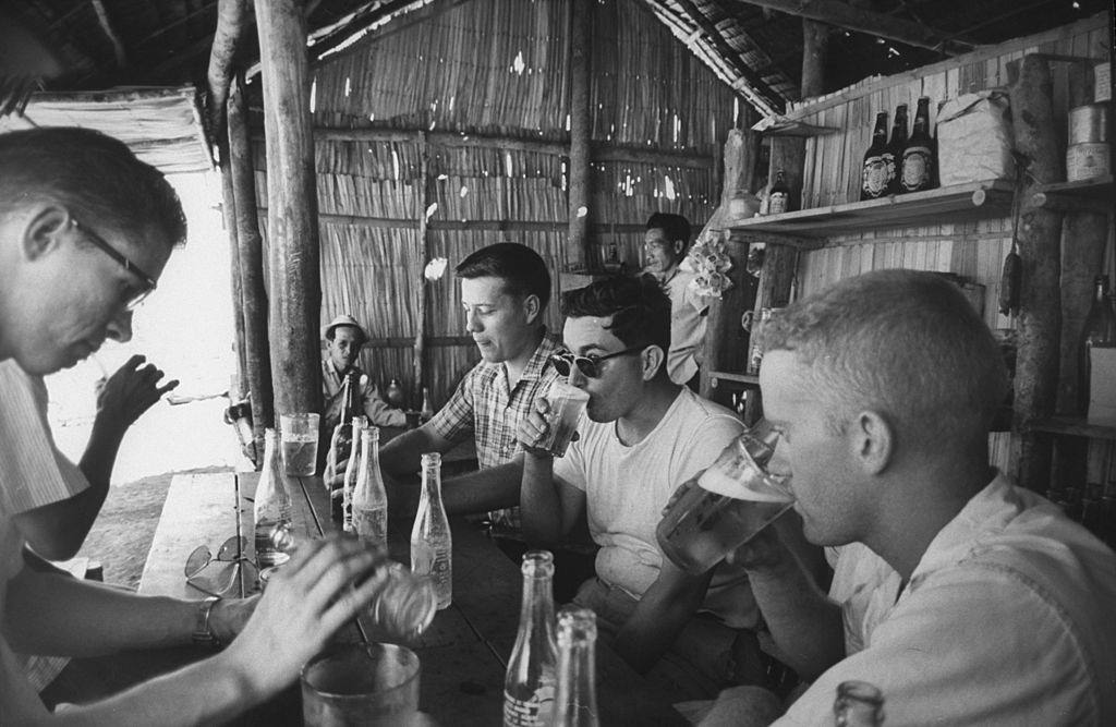 #19 Volunteers drinking in thatched shack, 1961.
