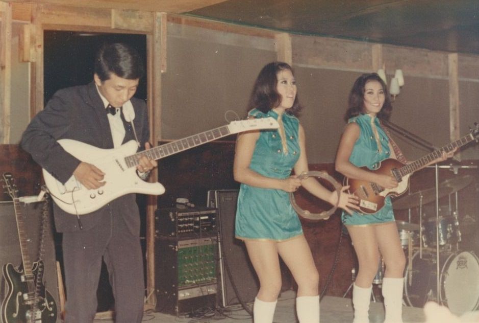 #62 A man playing a white electric guitar and two young women, wearing turquoise minidresses, playing the tambourine and another guitar, performing on a stage, Vietnam, 1965.