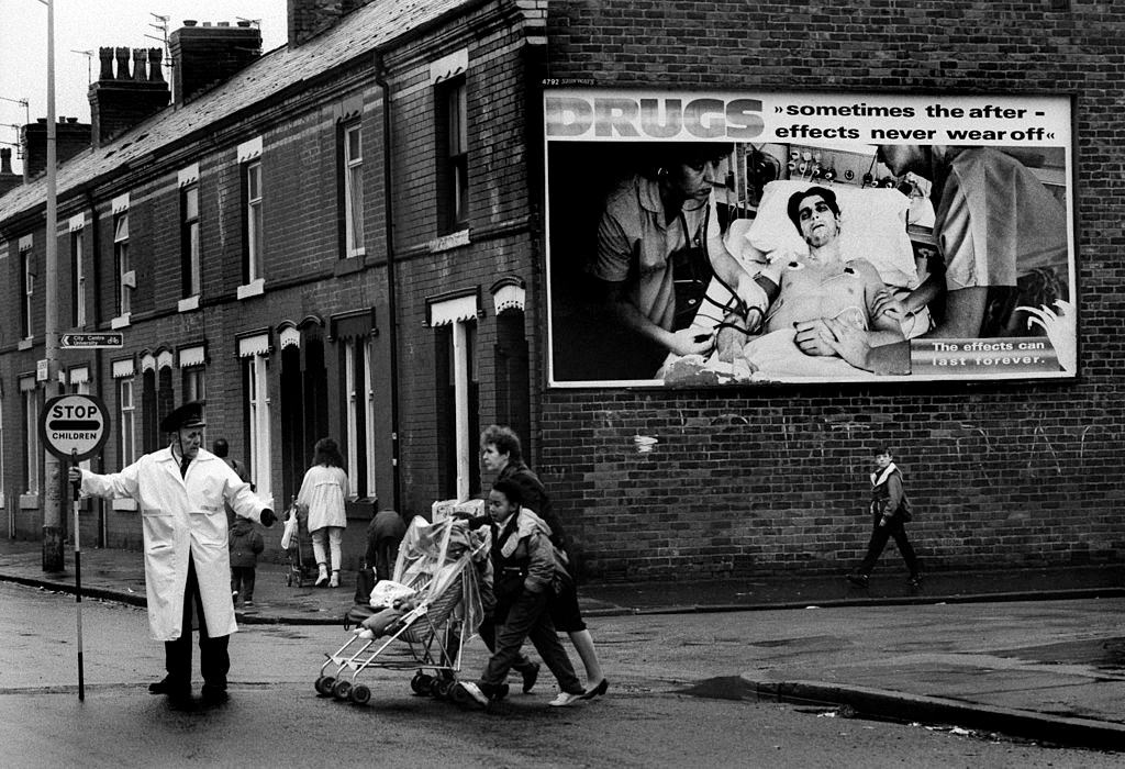 #32 A lollipop man guides children to school across a road in Moss Side, an inner city district of Manchester, 1990.