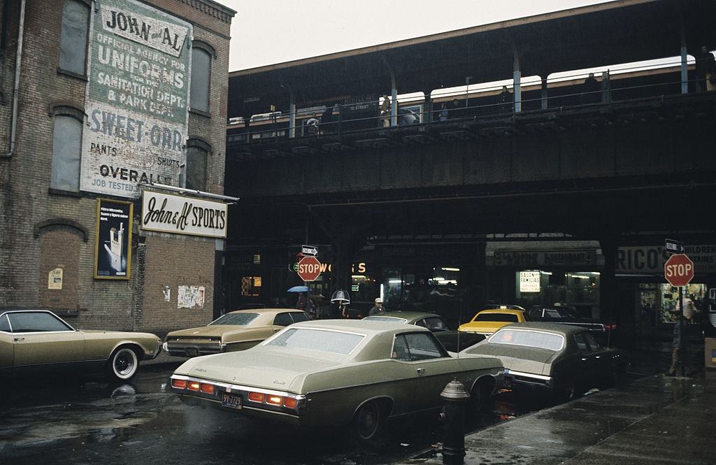 #20 A street with parked cars, passing under a subway traffic bridge, Brooklyn, 1973.