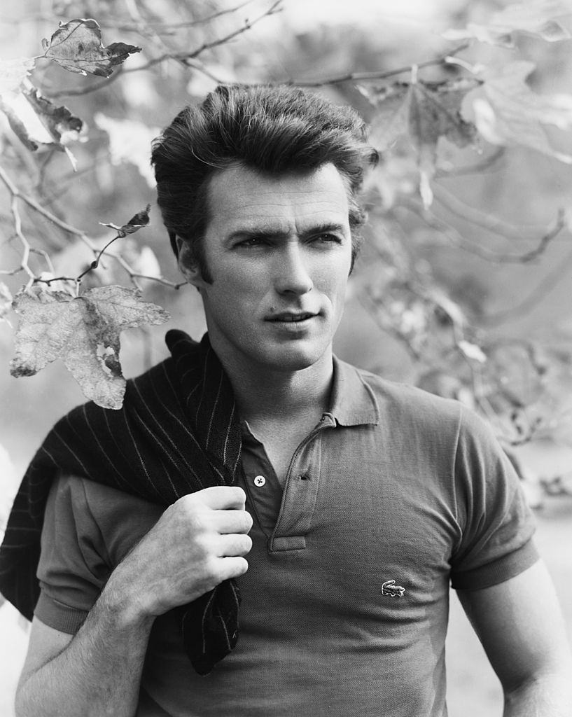 #18 Clint Eastwood squinting while posing under a tree, with a sweater slung over his shoulder, 1960.