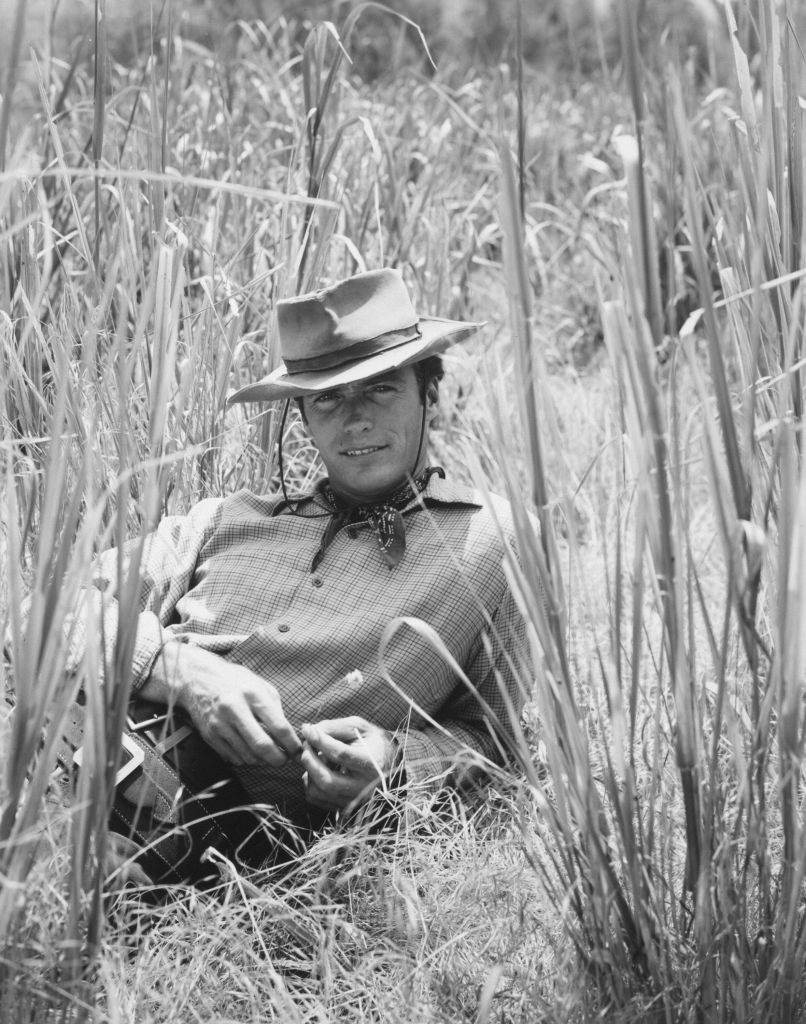 #33 Clint Eastwood smiles as he lies in tall grass for a portrait wearing a cowboy hat and Western outfit, 1961.