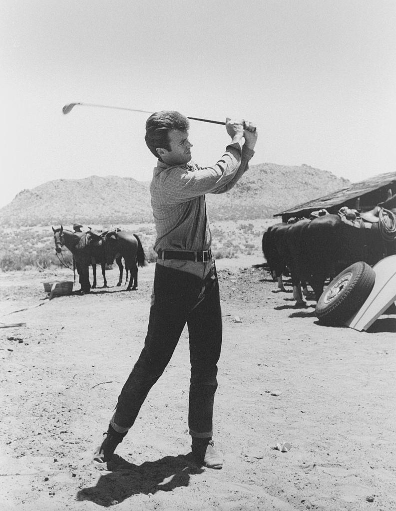 #41 Clint Eastwood wears chaps while he practices his golf swing as a horse looks on during a break in the shooting, 1962.