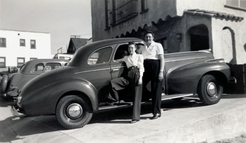 #10 Two ladies posing with a 1940 Oldsmobile Club Coupe in front of a house in Spanish Colonial style in bright sunlight, 1940