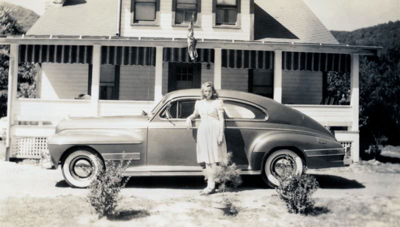 #11 A young lady in a white summer dress posing with a 1941 Oldsmobile Dynamic Cruiser Club Sedan in front of a white timber house, 1941