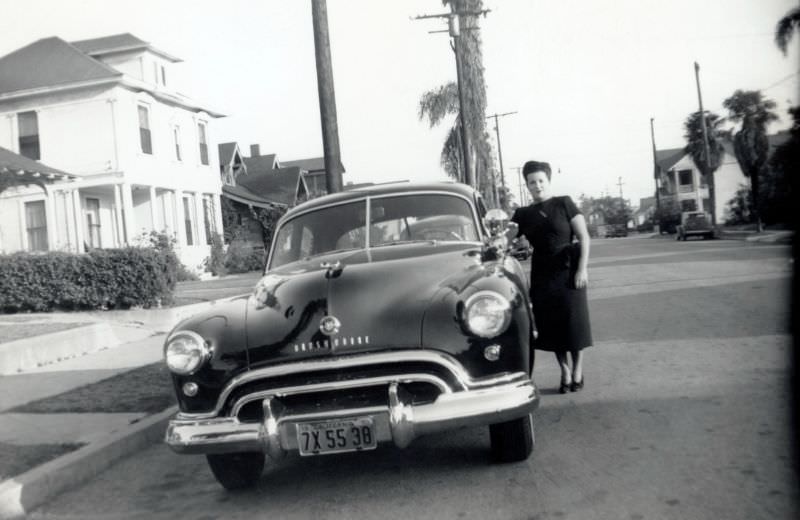 #13 A brunette lady posing with a 1948 Oldsmobile in a suburban street.