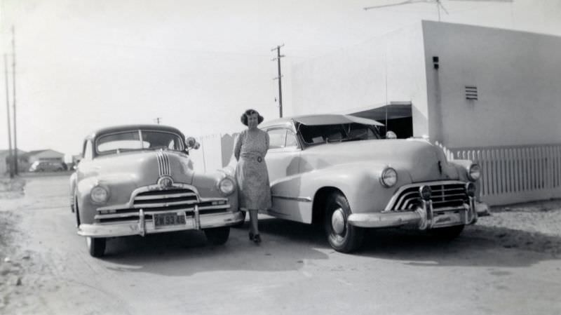 #15 A middle-aged lady posing with a pair of 1947 Pontiac and 1947 Oldsmobile in a dusty street on the outskirts of town.