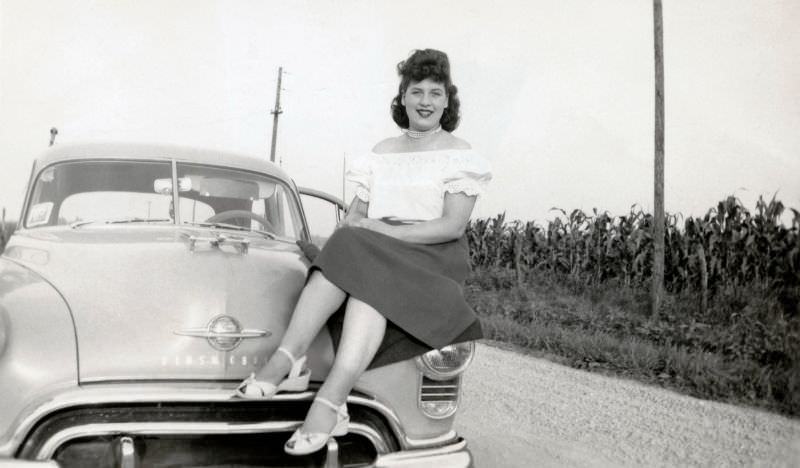 #17 A brunette lady posing on the fender of a 1949 Oldsmobile 88 on a gravel road in the cornfields, somewhere in rural America, 1950
