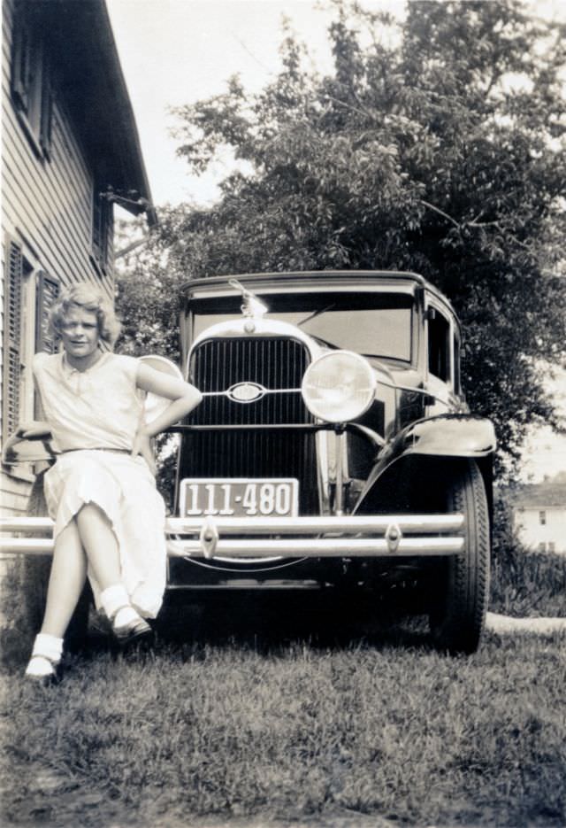 #2 A blonde lady in a white dress posing on the bumper of a 1930 Oldsmobile Six, adorned with an aftermarket propeller hood ornament,1930