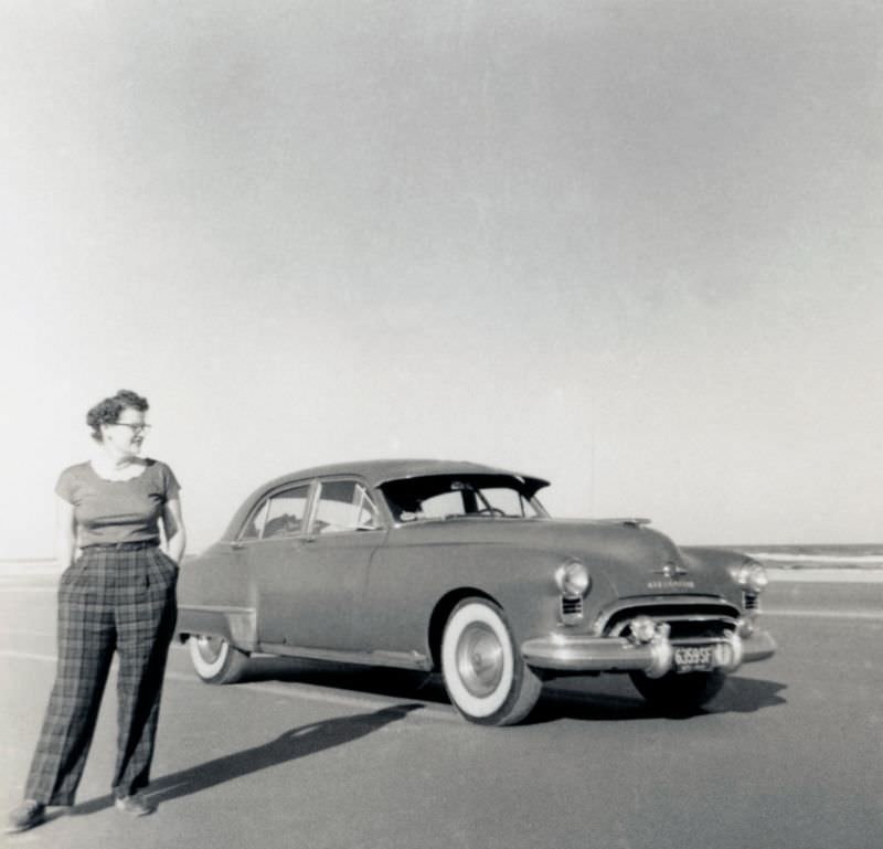 #20 A curly-haired lady dressed in tartan check trousers posing with a 1949 Oldsmobile 88 4-Door Sedan in late afternoon sunshine, 1952