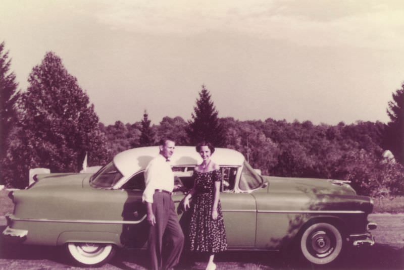 #22 A fellow in a white shirt and a lady in a floral dress posing with a green and white 1954 Oldsmobile Super 88 Holiday Coupe in the countryside.