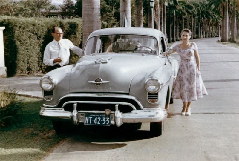 #25 A stylish lady in a summer dress and a fellow in a white shirt posing with a 1950 Oldsmobile 88 on an avenue lined with palm trees.