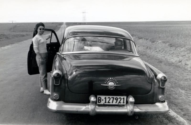 #26 A stylish young lady posing with a 1952 Oldsmobile 98 on the side of a long, straight road running through a plain landscape