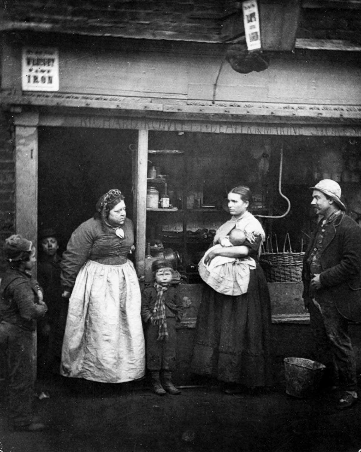 #24 People in front of a rag shop in Lambeth, London, where the Thames annual tidal overflow causes hardship to the locals, 1877.