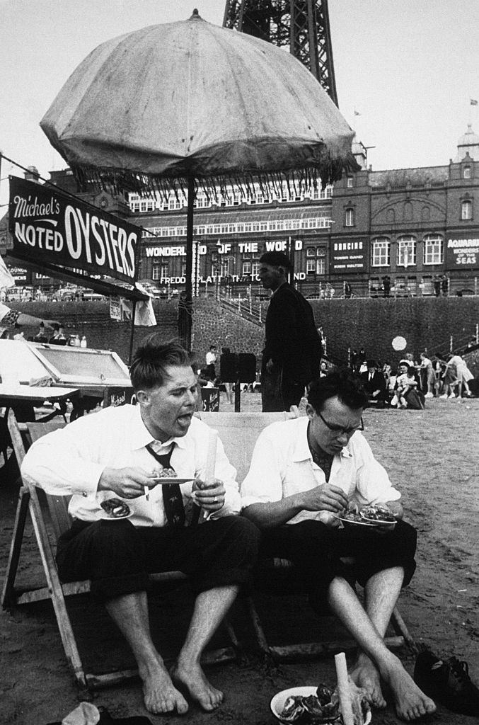 #70 The comedians Eric Morecambe and Ernie Wise, enjoying some of the local delicacies on the beach at the foot of the Blackpool Tower, 1953.