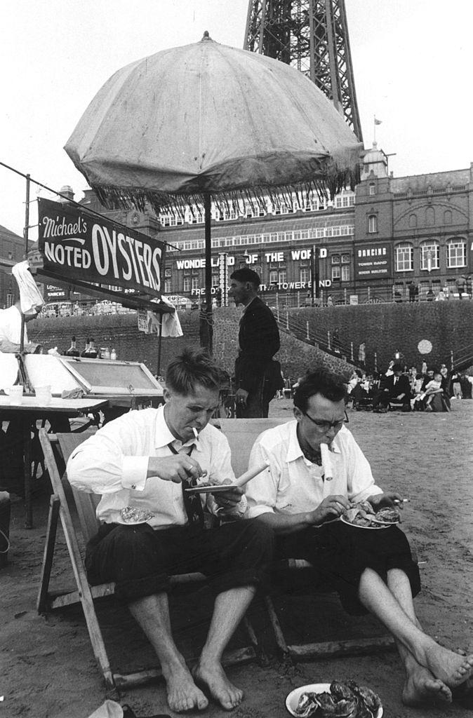 #21 British comedy double act Eric Morecambe and Ernie Wise, in rolled up tousers, eating seafood on the beach at Blackpool, 1953.
