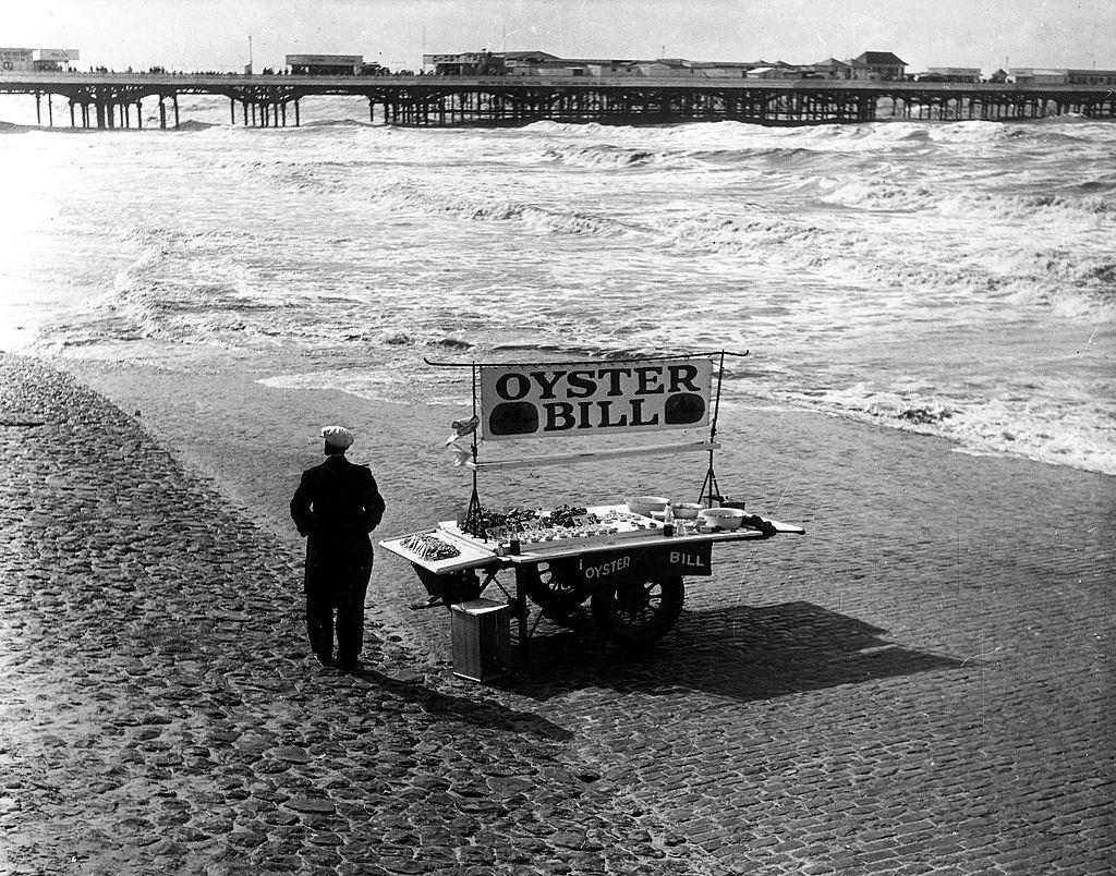 #38 A man standing with a barrow displaying “Oyster Bill” on the sand at Blackpool, 1951.