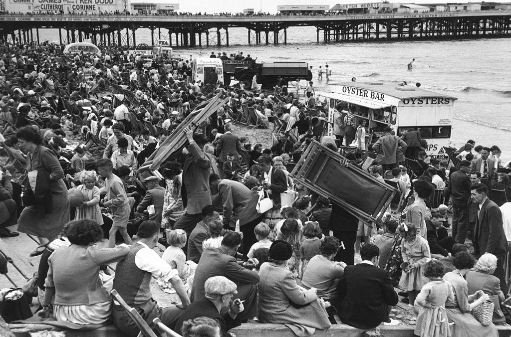 #58 A crowd of people on the beach at Blackpool, some carrying deckchairs, July 1956.
