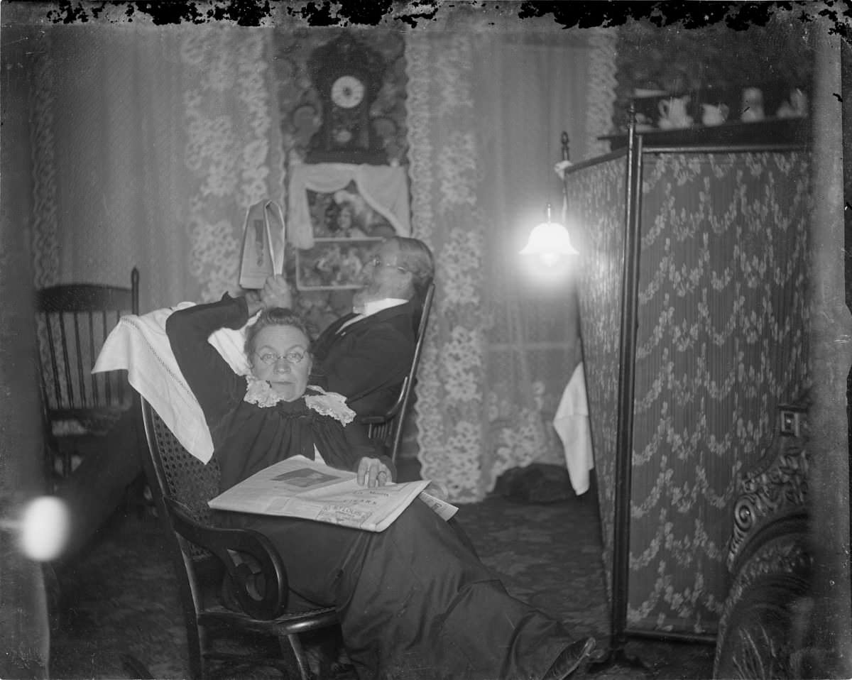 #14 Woman sitting in rocking chair with newspaper on lap and man seated behind her – George Silas Duntley Photographs 1899-1918