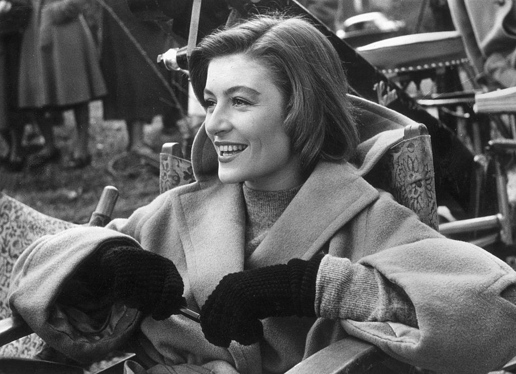 #8 Anouk Aimee, wearing a camel-hair overcoat and gloves, laughs while sitting in a folding chair on a film set, 1955.