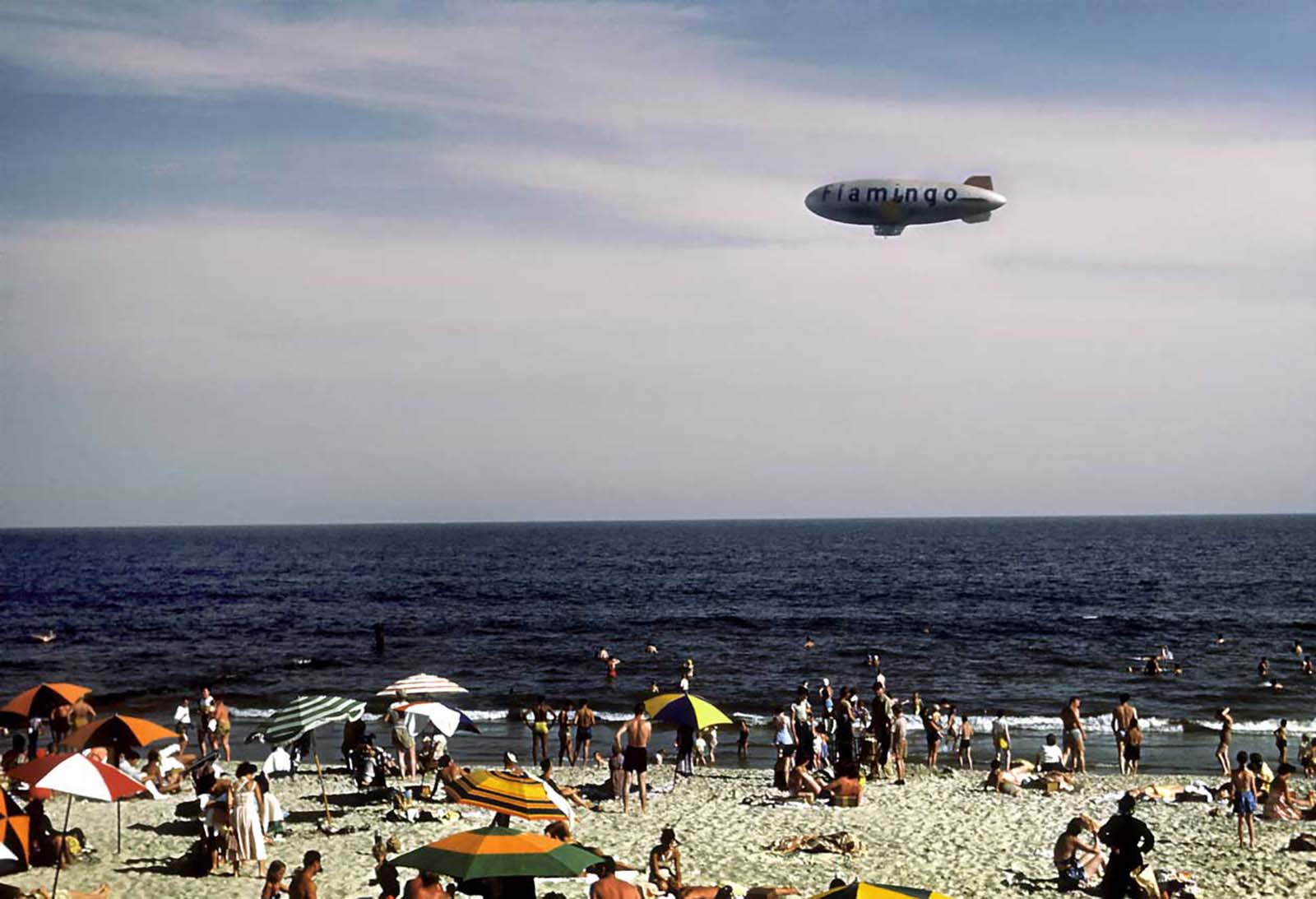 #1 Sunbathers and swimmers on the beach while a blimp with the word Flamingo on the side glides above.