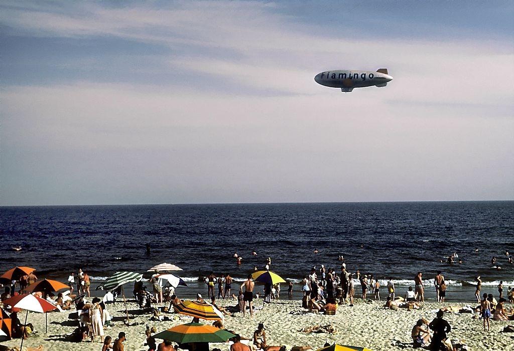 #14 Sunbathers and swimmers frolic on Coney Island beach while a blimp with the word Flamingo on the side glides above, 1948.