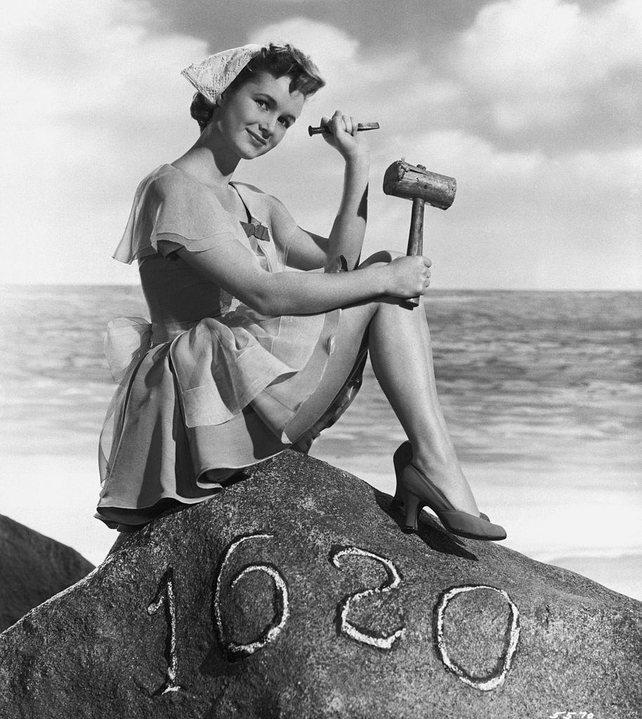 #38 Debbie Reynolds, dressed as a pilgrim, sits on top of a replica of Plymouth Rock holding a mallet and a chisel.