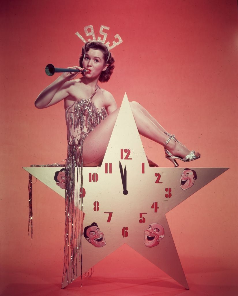 #40 Debbie Reynolds, wearing a 1953 tiara and a costume with tinsel, blows a horn while sitting atop a star-shaped clock in a promotional portrait for New Year’s Eve.