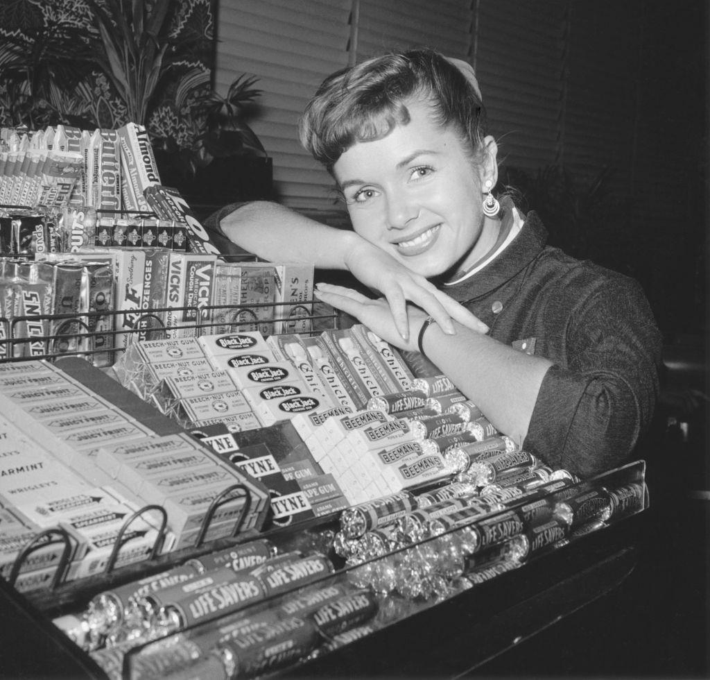 #41 Debbie Reynolds smiles while leaning upon a counter filled with a display of various candies, 1952.
