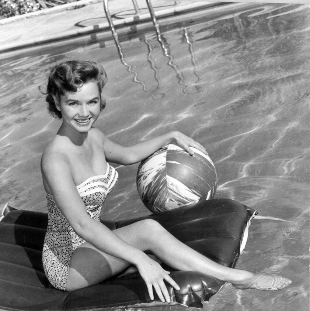 #50 Debbie Reynolds, wearing a strapless one-piece swimsuit, holding a beach ball while sitting on an air mattress in a swimming pool, 1955.