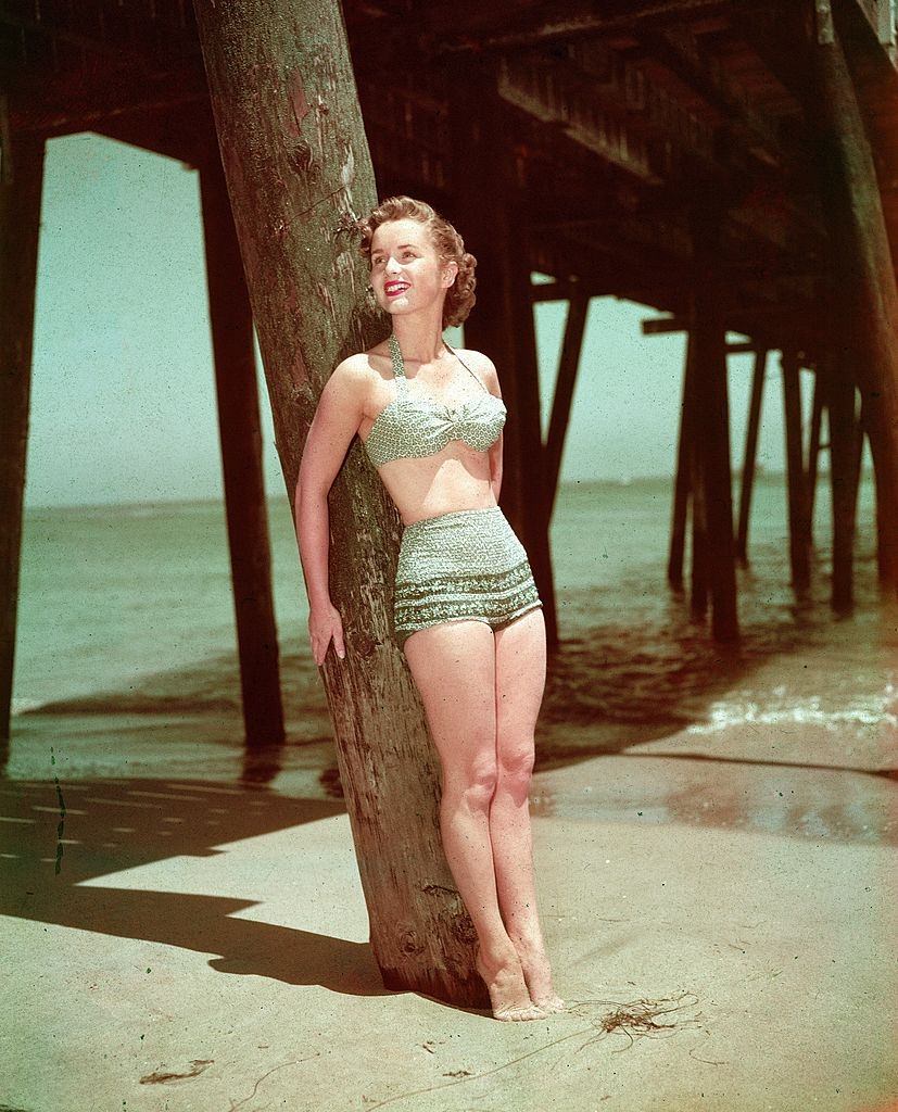 #13 Debbie Reynolds wearing a two-piece swimsuit while leaning against a wooden pier post on a beach, 1950s.