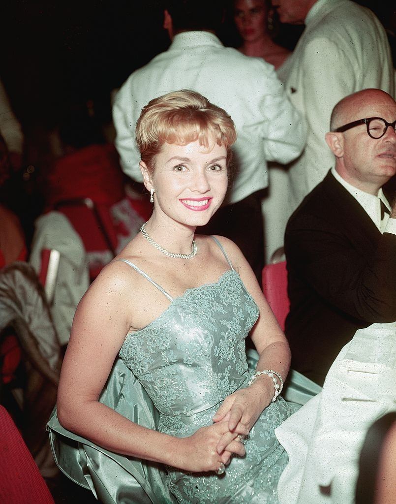 #55 Debbie Reynolds, wearing an evening dress, smiles while sitting in the crowd at a formal event, 1950s.