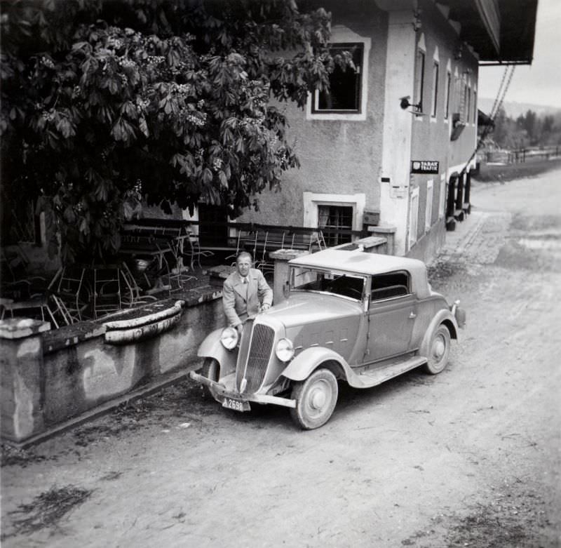 #7 A fellow in a light-coloured raincoat posing with a Renault Monaquatre Cabriolet-Spider convertible in front of a rural inn.
