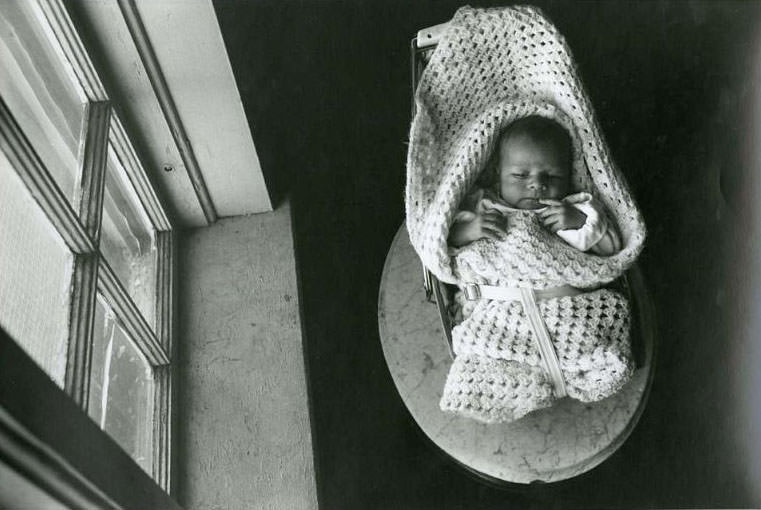 #13 A newborn wrapped in a white blanket, lying in a baby chair, which is on top of a small oval, marble table and net to a window, Jefferson Park, 1975