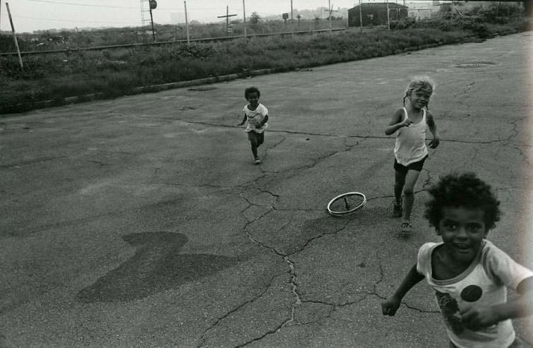 #15 Three children running along a road past a bicycle wheel with spokes lying on the ground. The railroad tracks are visible in the background, Jefferson Park, 1975