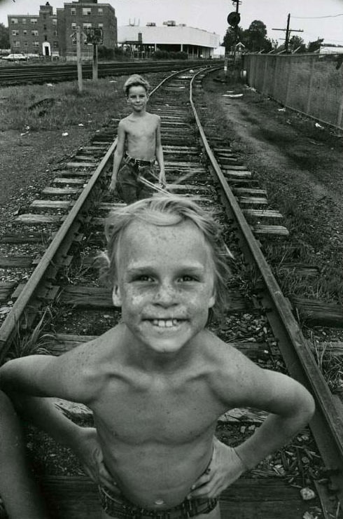 #8 Two boys standing on railroad tracks, both are shirtless. The Jefferson Park Housing Project is visible in the background, Jefferson Park, 1973