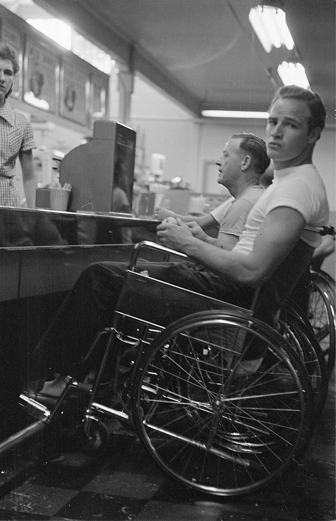 #46 Marlon Brando sitting in a wheelchair at a coffee shop counter, during the filming of “The Men”.