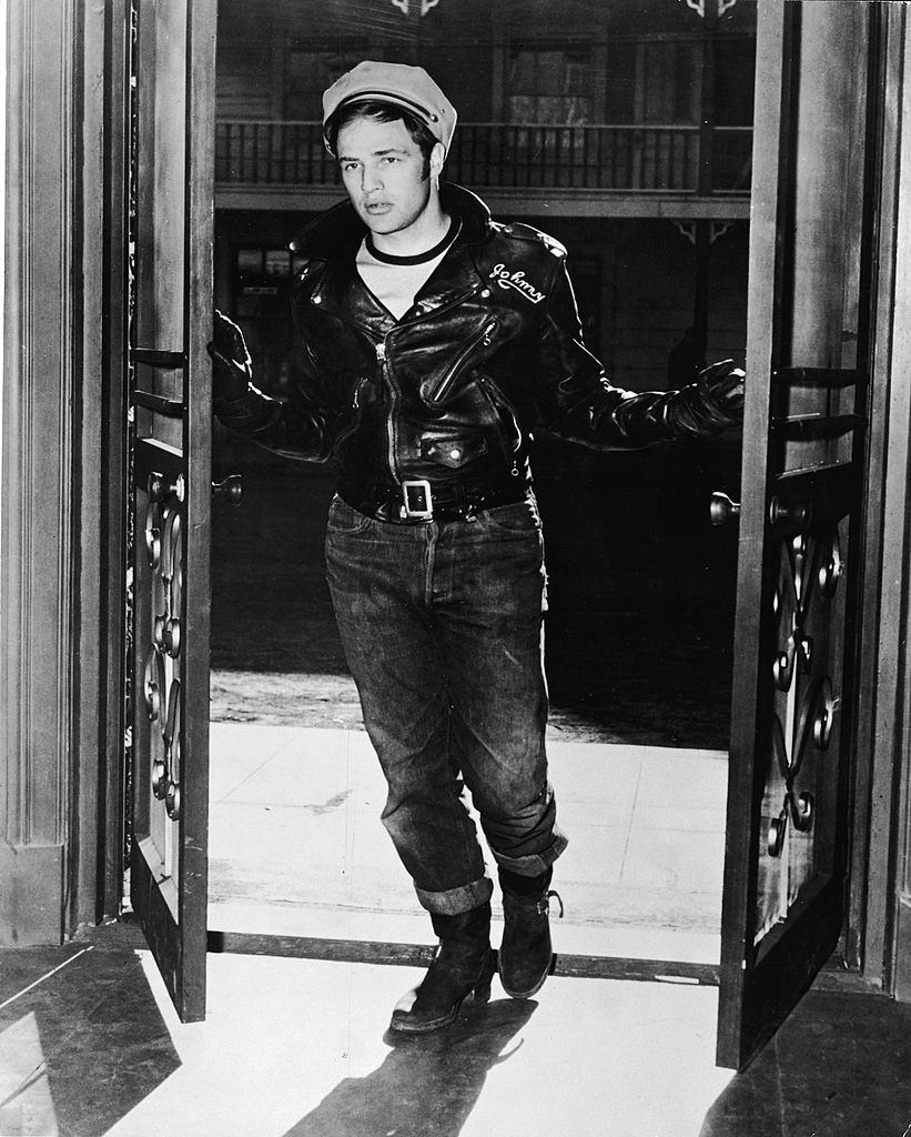 #62 Marlon Brando stands in an open doorway, wearing a leather jacket and hat, 1953.