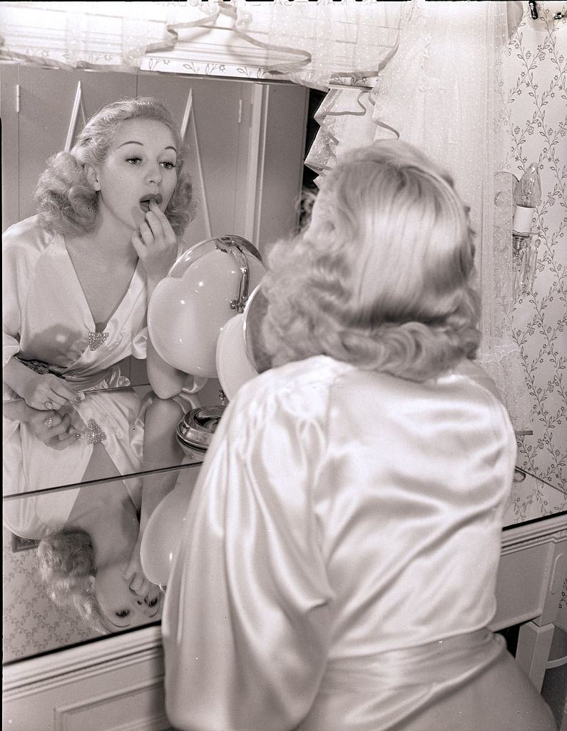 #27 Bertty Grable, wearing a satin robe and applying lipstick in a mirror, 1939.
