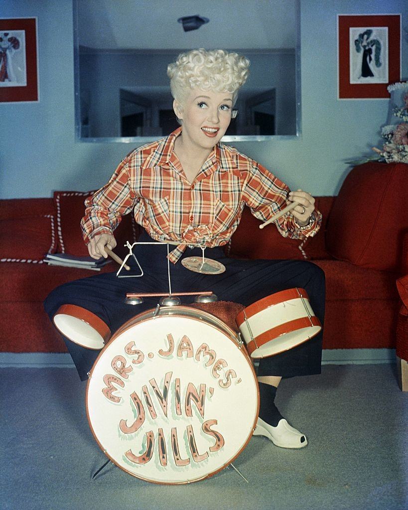 #34 Betty Grable wearing a plaid shirt, sitting behind a child’s drum kit, which has ‘Mrs James Jivin’ Jills’, 1940.