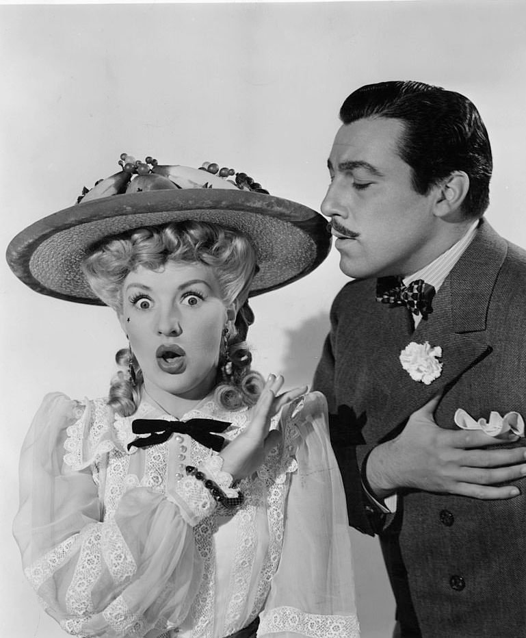 #71 Betty Grable puts her hand up as Cesar Romero holds his heart in a scene from the film ‘Coney Island’, 1943.