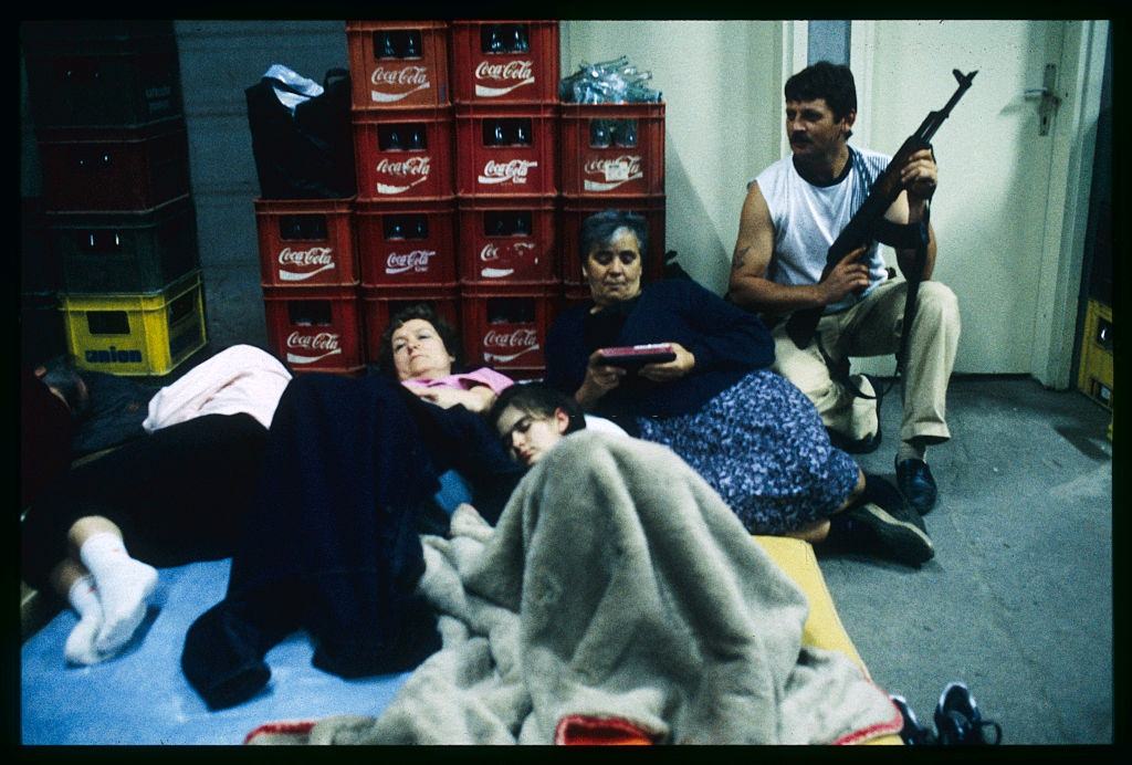 #13 People lie on beds and sit on the floor while a man holds a rifle in a shelter during bombings in Osijek.