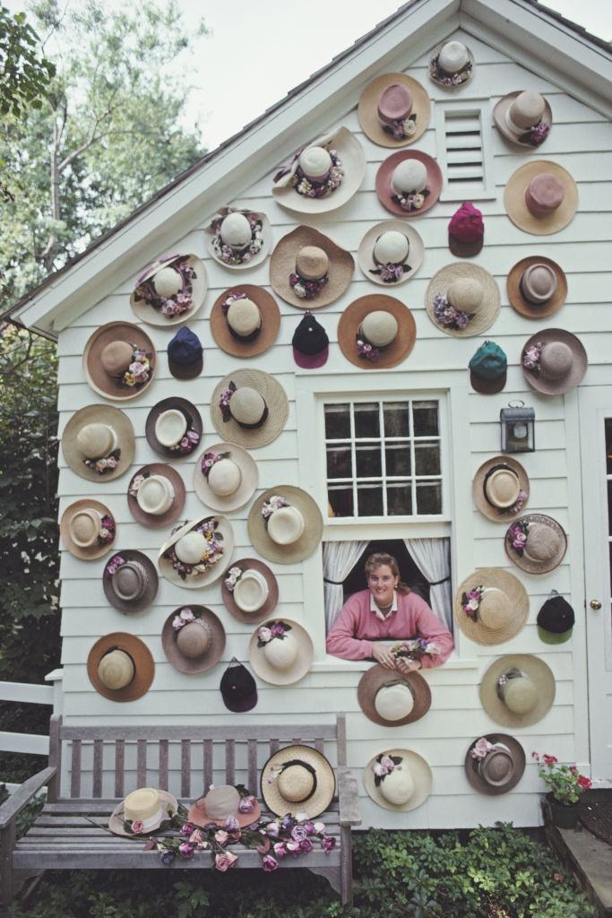 #21 Hat designer Darcy Creech appearing from the window of a house with its facade covered with hats, September 1991