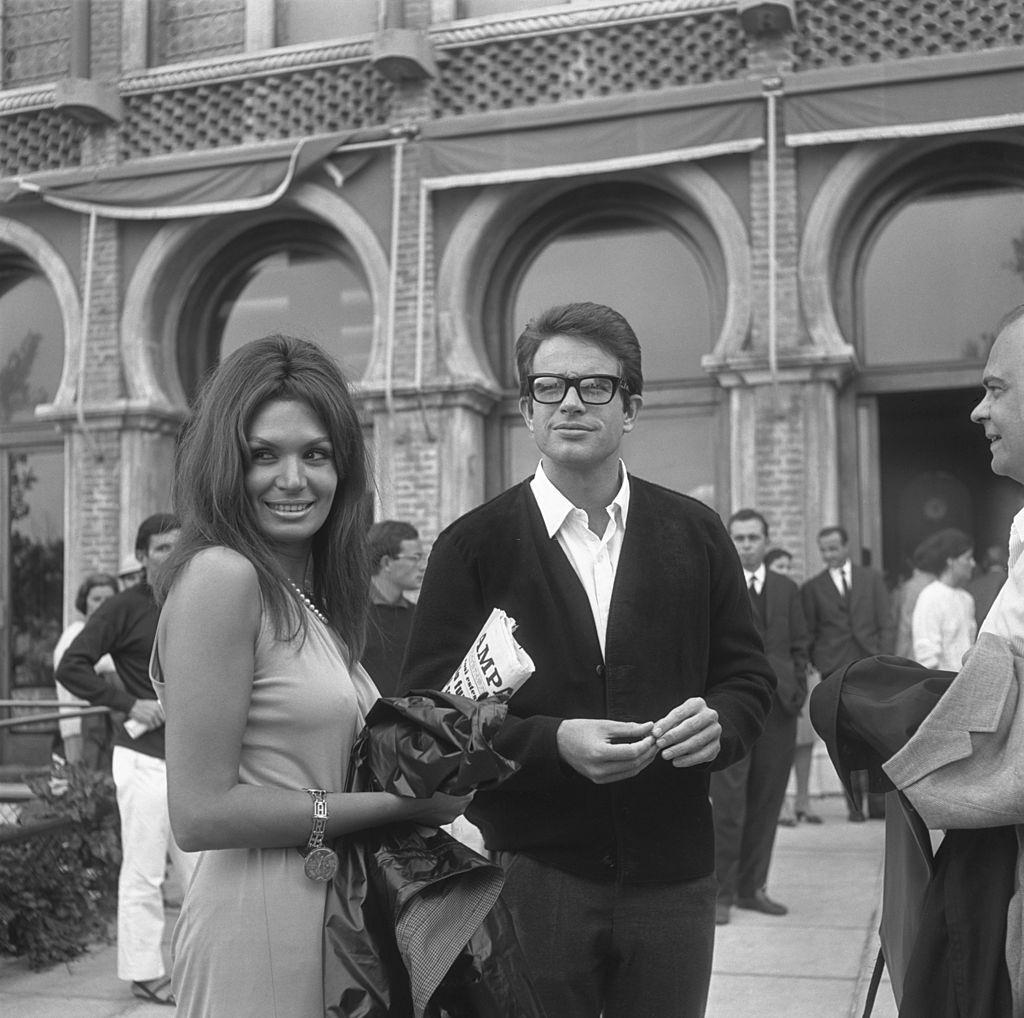 #37 Warren Beatty, wearing a cardigan and glasses, portrayed outside the Excelsior Hotel while standing next to a woman holding a newspaper, 1965.