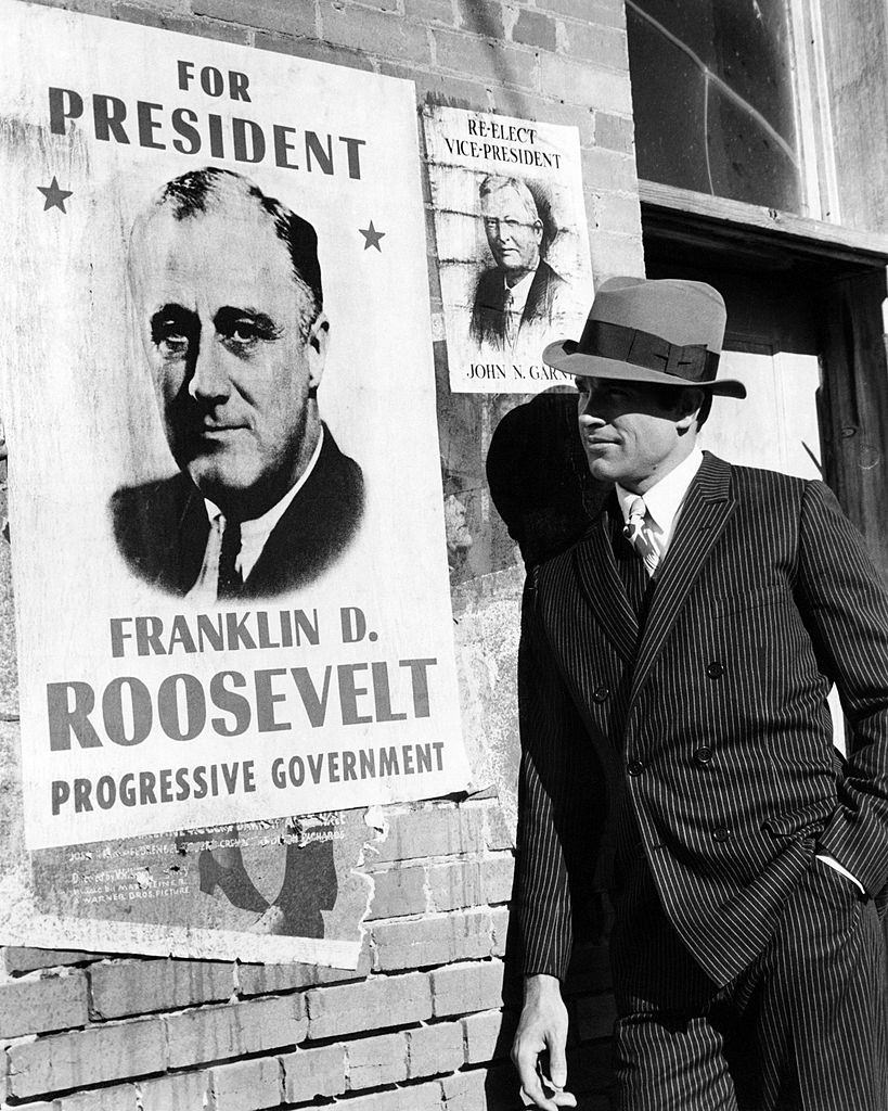 #53 Warren Beatty, as Clyde Barrow, posing next to a Roosevelt campaign poster in a promotional portrait for ‘Bonnie And Clyde’, 1967.