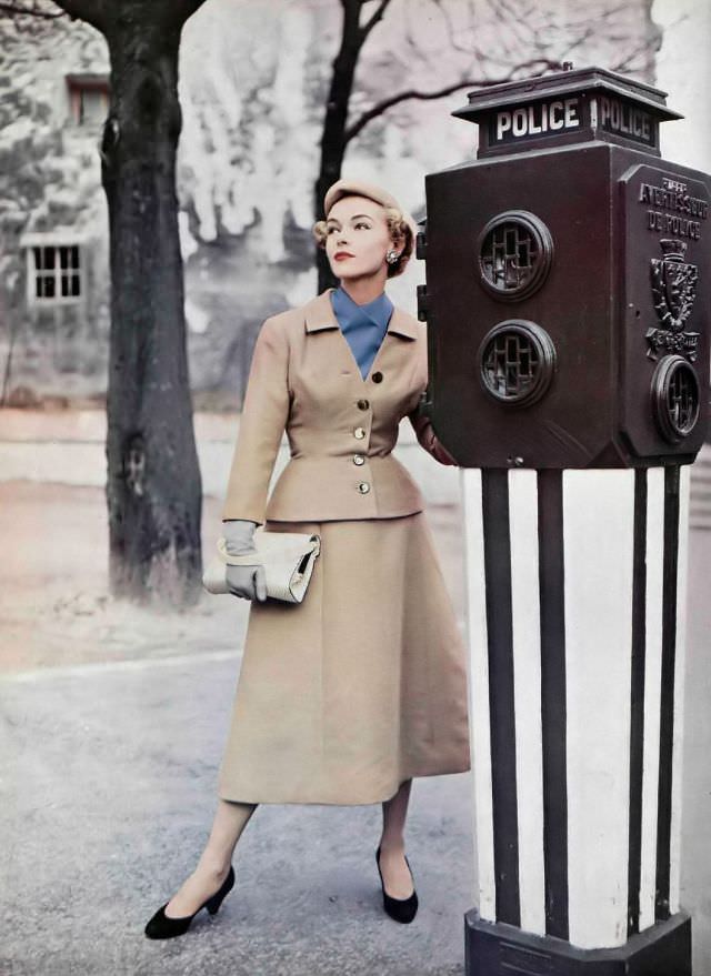 #21 Joan Olson in classic beige suit, the skirt is bell-shaped, jacket has nipped waist and low neck that shows a bright blue scarf by Jean Patou, 1954
