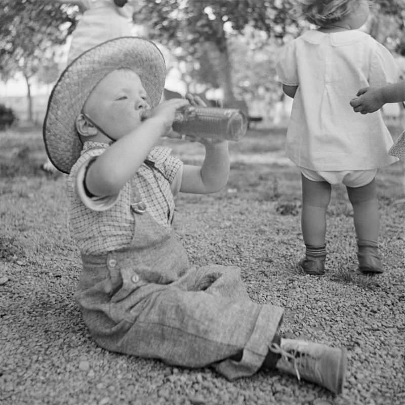 #30 Little boy sitting on the ground, wearing a hat and drinking from a bottle